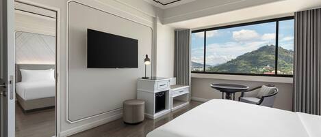 A modern hotel room with a large bed, a flat-screen TV, a desk with a chair, and a view of a mountainous landscape through a large window.