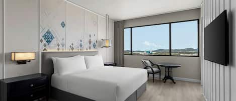 A modern hotel room with a large bed, a desk, a chair, a TV, and a view of the city through large windows.