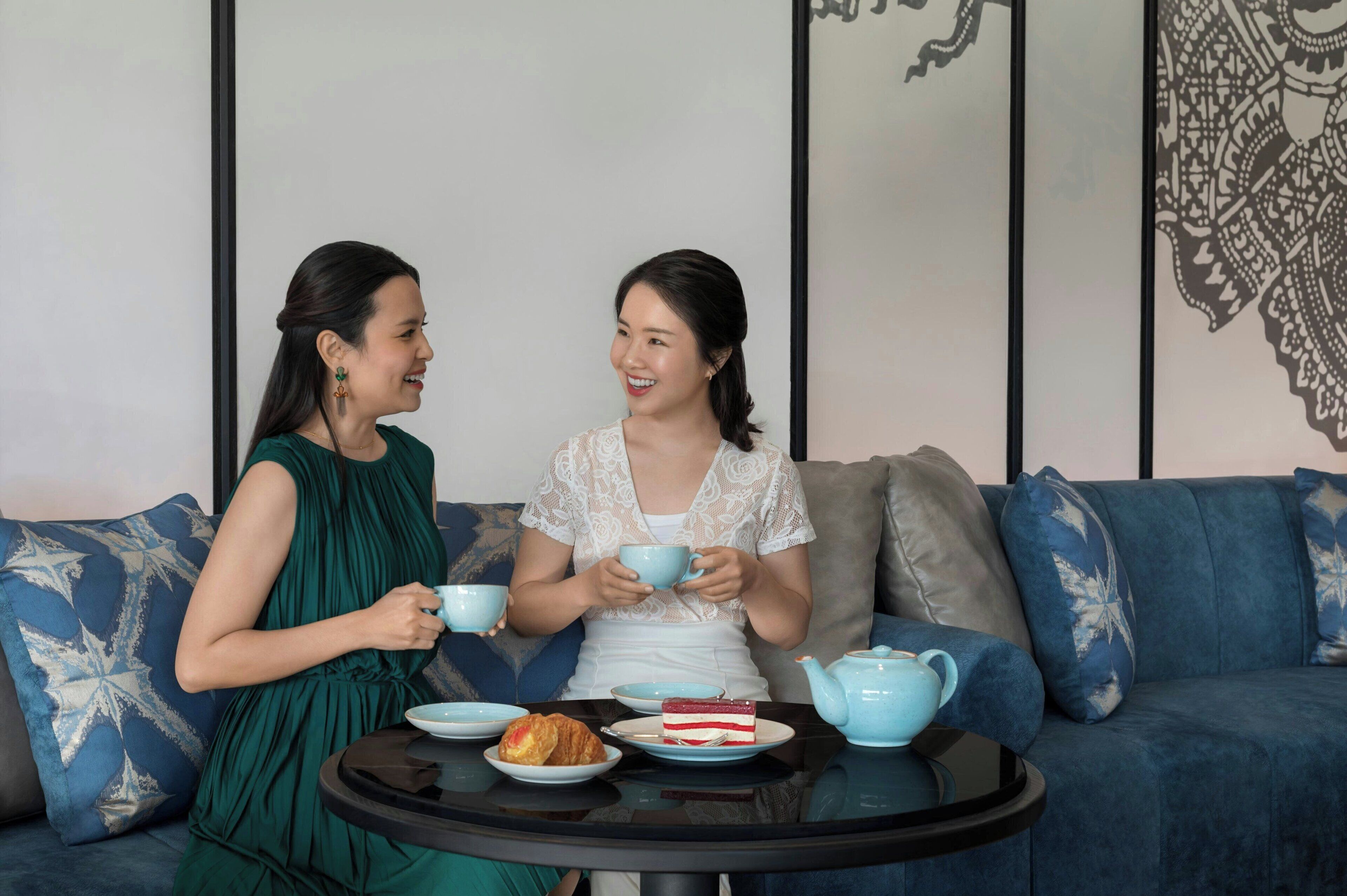 Two people sitting on a sofa with a table between them. The table has a teapot, cups, and a cake.