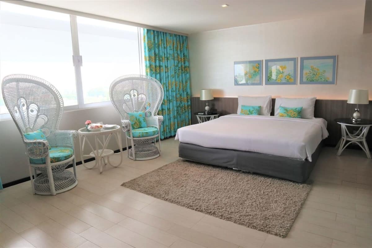 A hotel room with a large bed, two white wicker chairs, a small round table, and a view of the outdoors through a window with blue curtains.