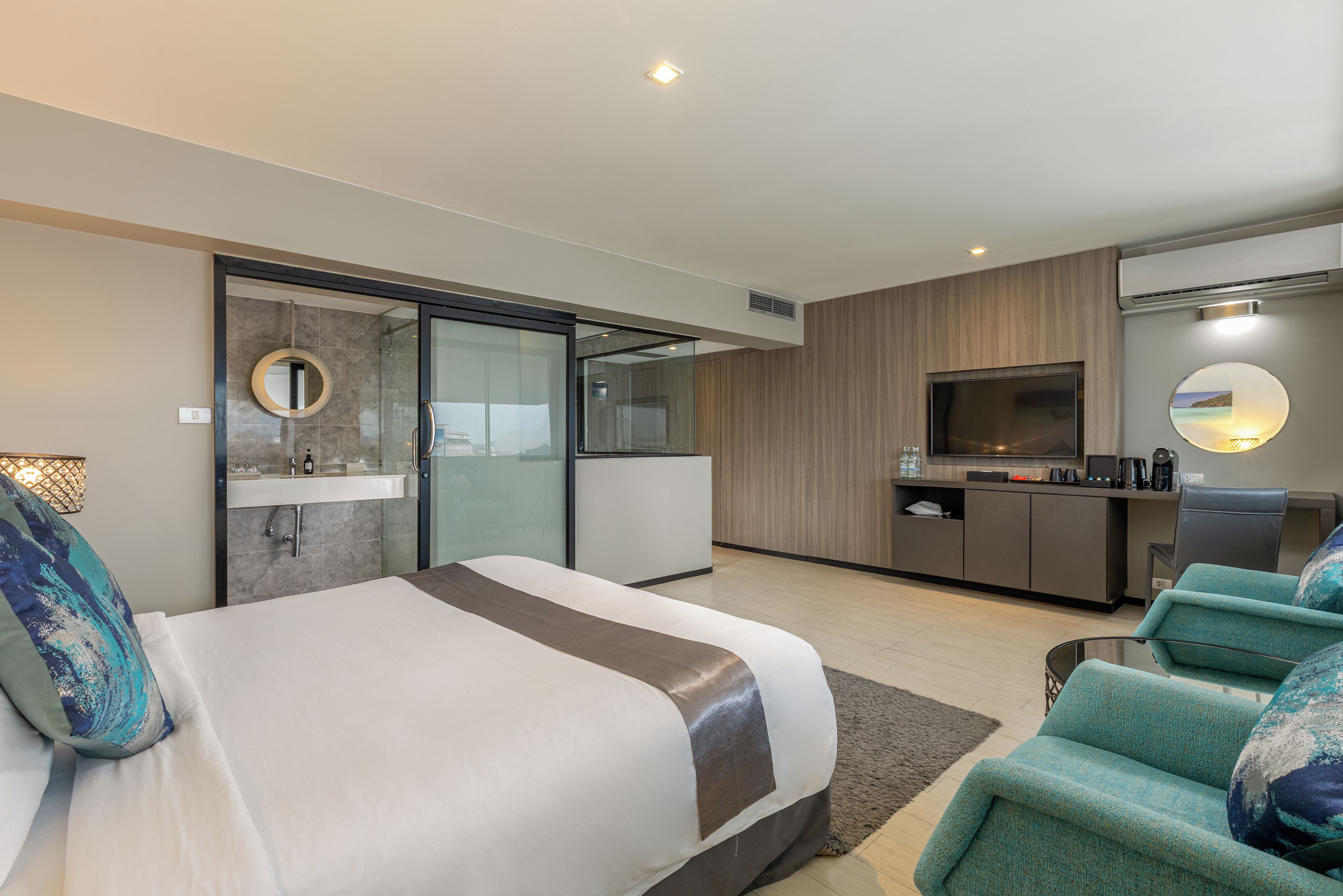 A modern hotel room with a bed, seating area, television, and a bathroom with a glass shower enclosure.