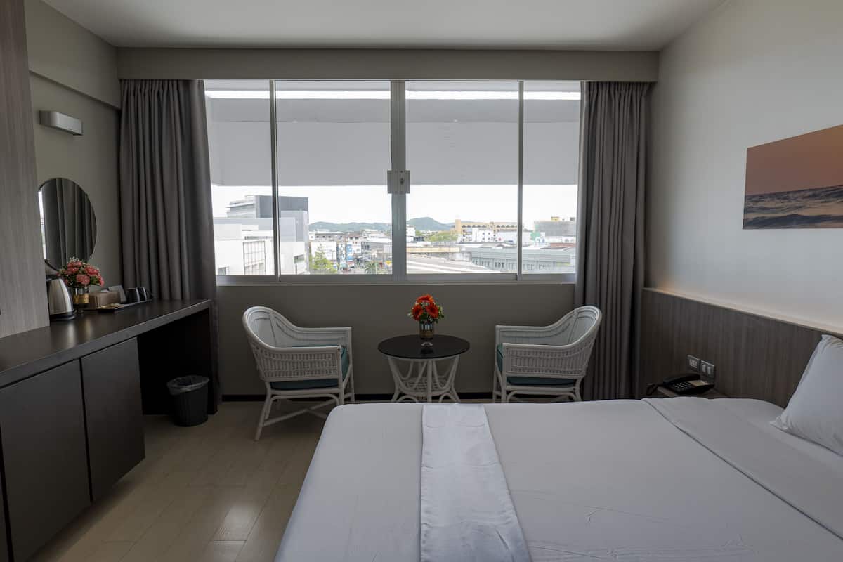 A hotel room with a large bed, a desk, two chairs, a small table with flowers, and a view of the city through large windows.