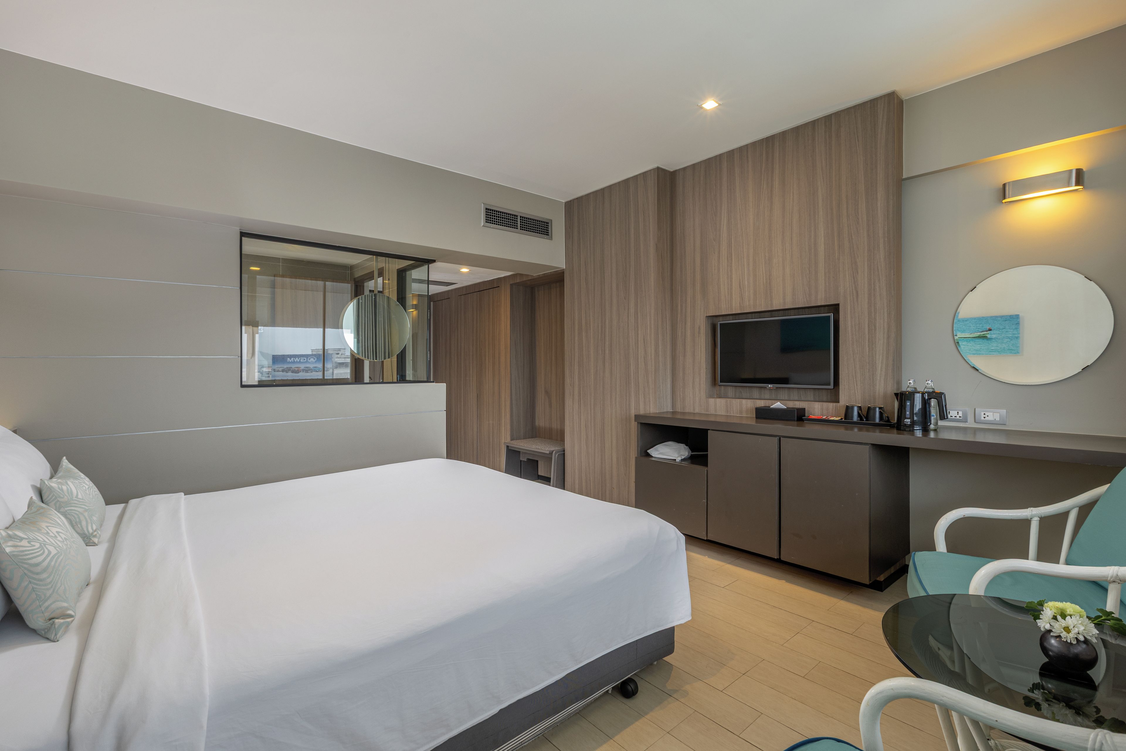 A hotel room with a bed, a desk with a flat-screen TV, a mirror, and a small table with a vase.