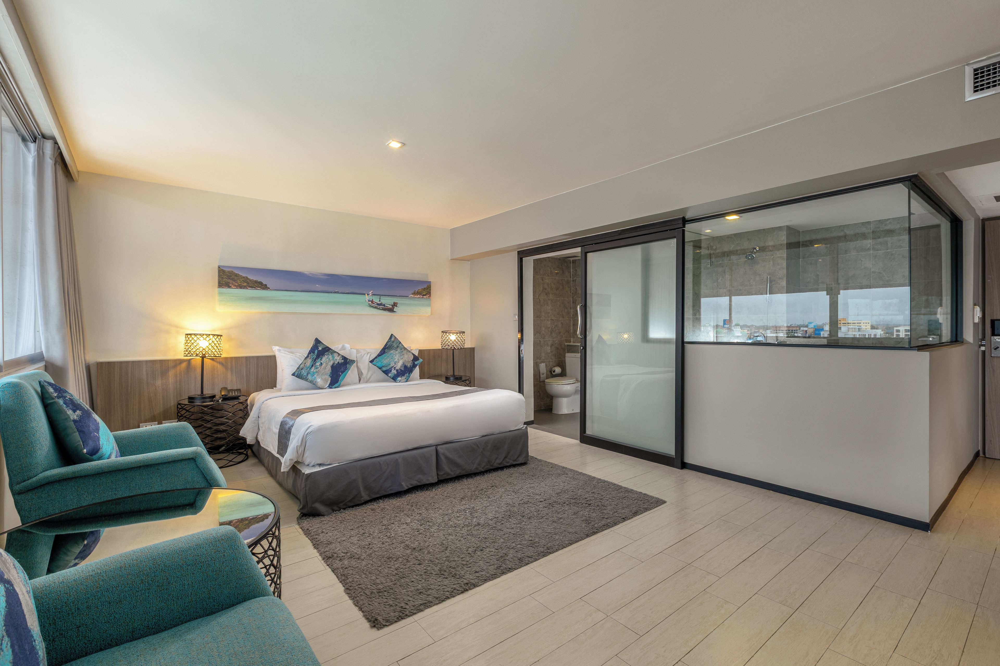 Habitación de hotel moderna con cama, zona de estar con sillas, ventana grande con vista a la ciudad y baño con puerta de cristal.