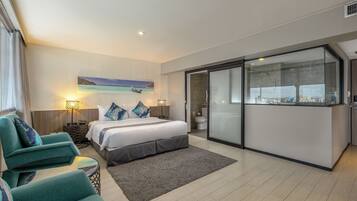 Habitación de hotel moderna con cama, zona de estar con sillas, ventana grande con vista a la ciudad y baño con puerta de cristal.