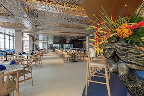A restaurant with multiple tables and chairs, a decorative wall with a sculpture, and a view of the outside through large windows. - Pearl Hotel Phuket (Phuket)