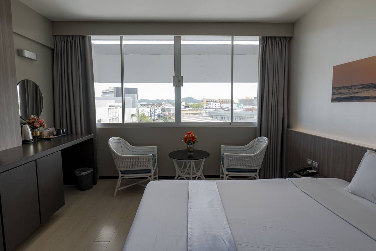A hotel room with a bed, a desk, two chairs, a small table with flowers, a view of the city, and a painting on the wall.