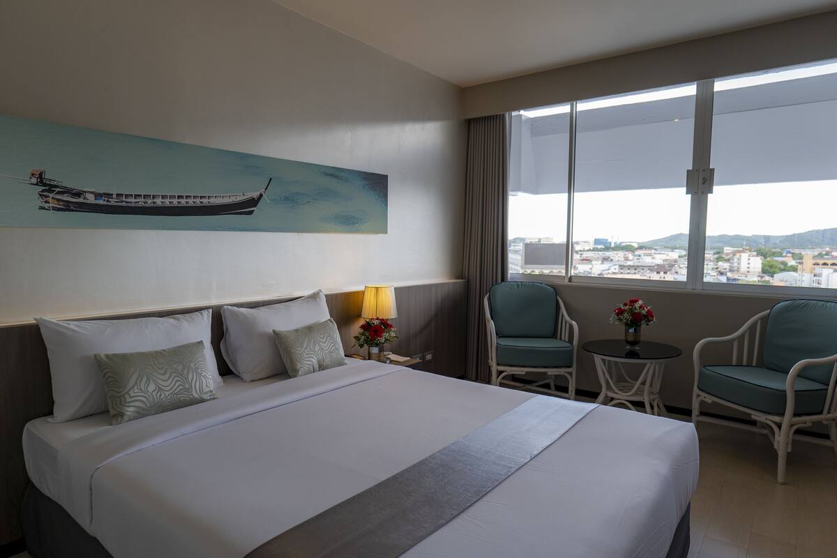 A hotel room with a large bed, two chairs, a small table, a lamp, and a painting of a boat on the wall.