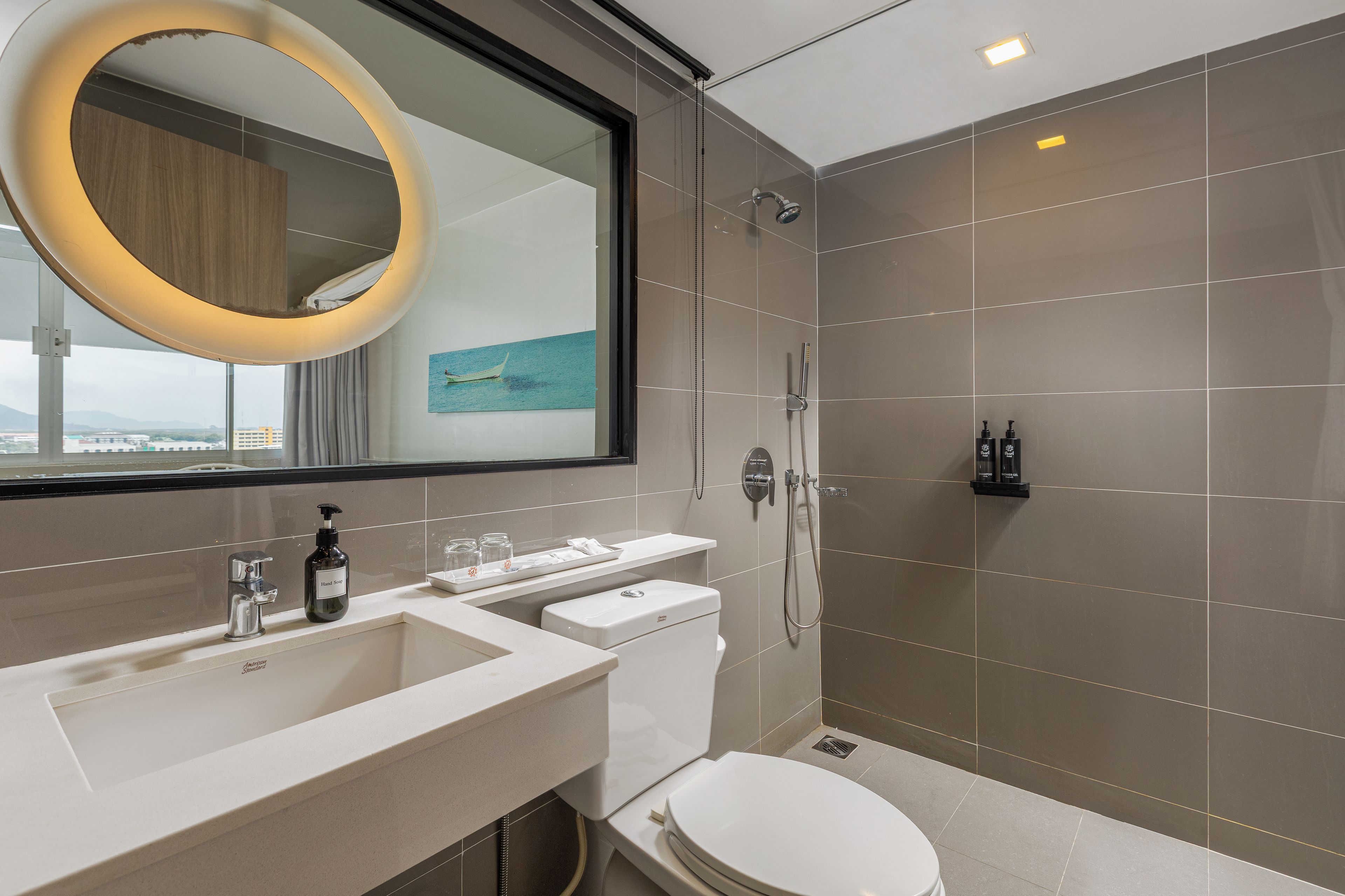 Un baño moderno con un espejo grande, ducha y retrete. Hay un lavabo con dispensador de jabón y una bandeja con vasos.
