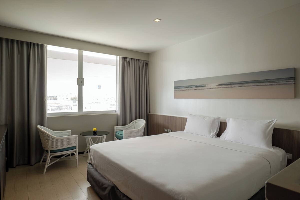 A hotel room with a large bed, two chairs, a small table, and a window with curtains.