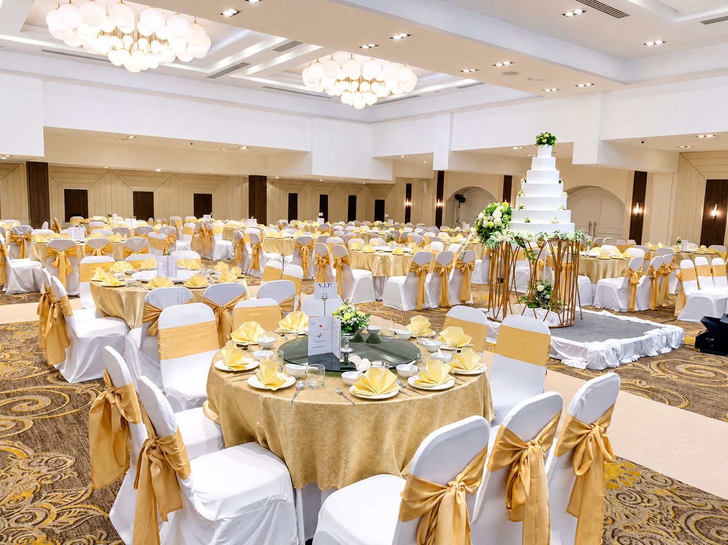Un salón de banquetes con mesas redondas, sillas con cintas doradas y una torta blanca de varios pisos como centro de mesa.