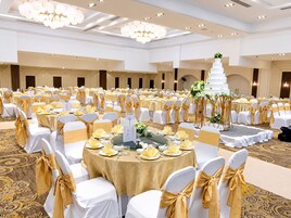 Un salón de banquetes con mesas redondas, sillas con cintas doradas y una torta blanca de varios pisos como centro de mesa.