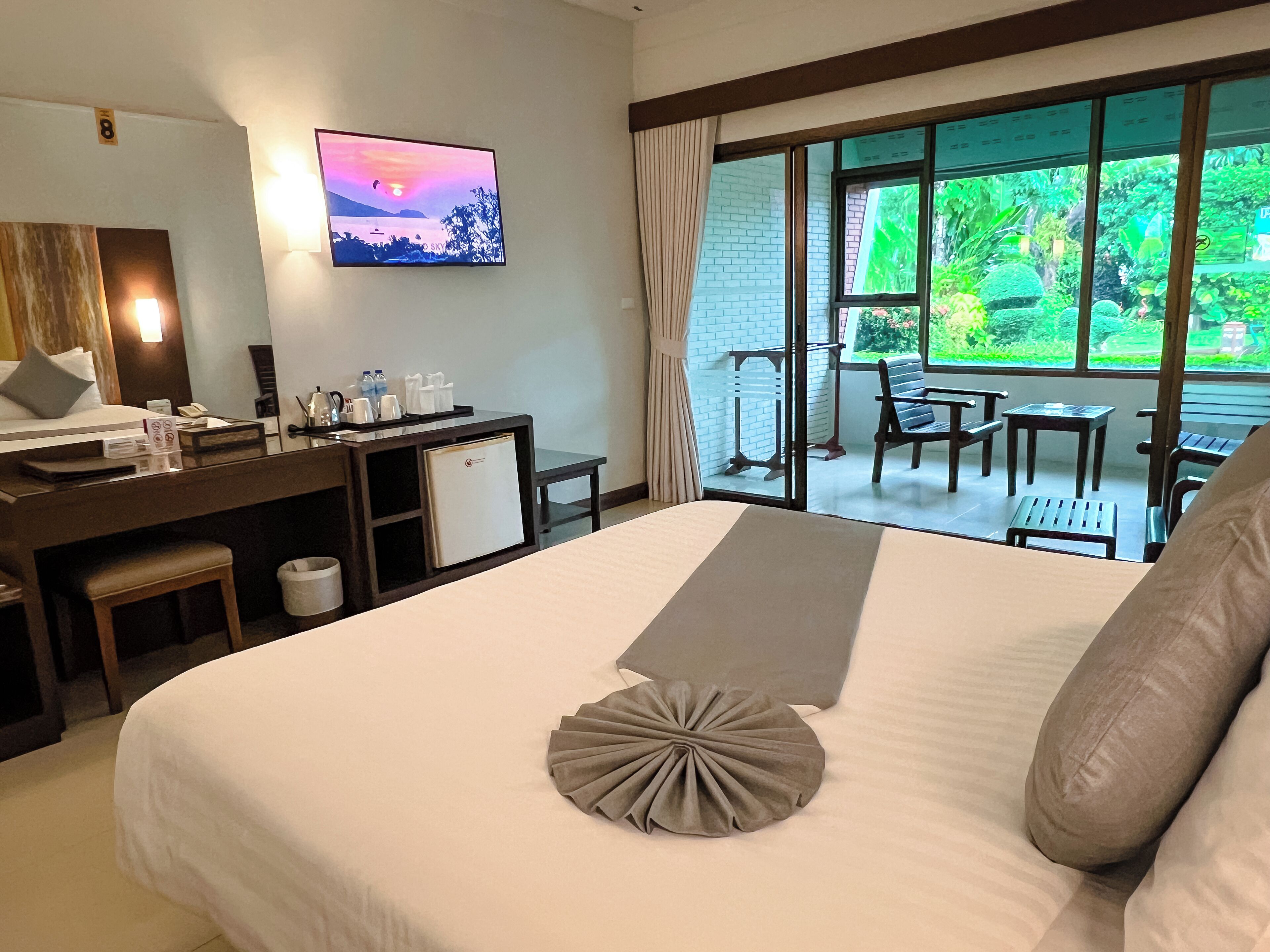 Habitación de hotel con cama, escritorio con computadora, refrigerador pequeño y televisión en la pared. Incluye balcón con sillas y mesa, y vista a zonas verdes.