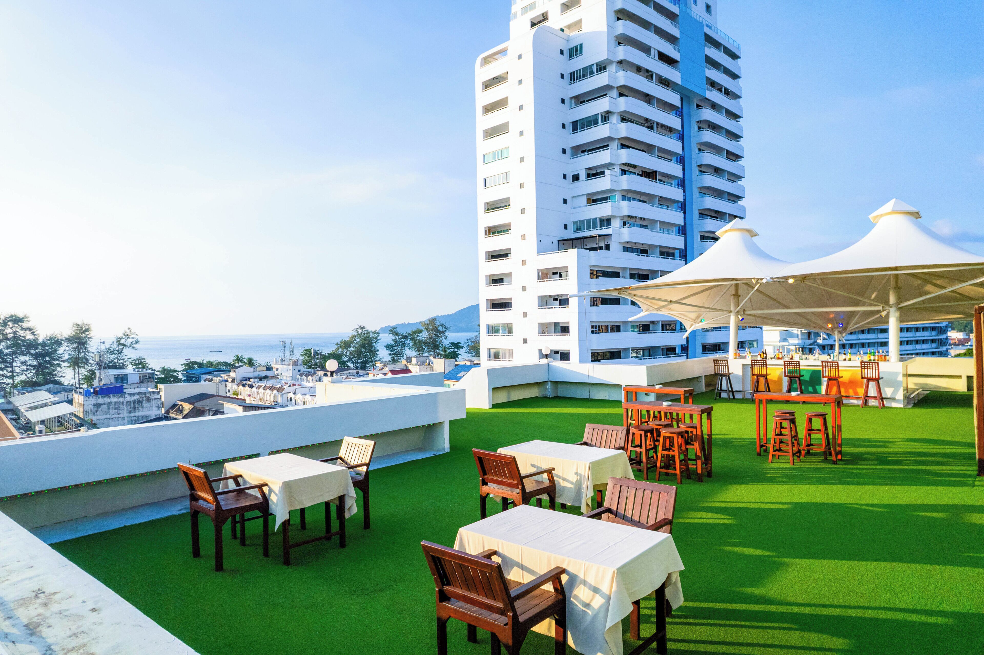 A rooftop terrace with tables and chairs, a bar area, and a view of the sea and buildings.
