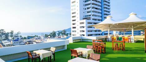 A rooftop terrace with tables and chairs, a bar area, and a view of the sea and buildings.