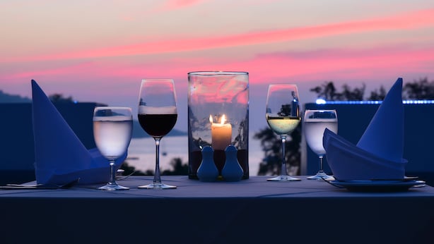 A table set with four glasses of wine, a lit candle, and two salt and pepper shakers, set against a sunset backdrop.