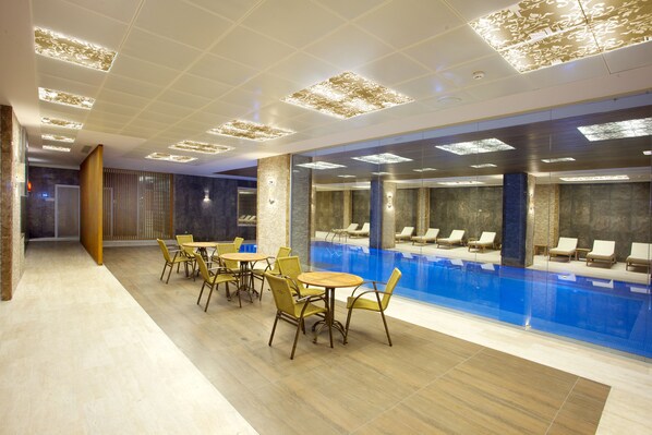 A modern indoor pool area with lounge chairs and tables, a clear pool, and recessed lighting on the ceiling.