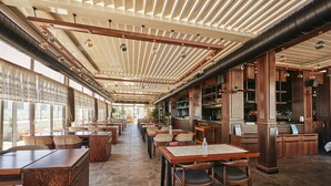 A spacious restaurant with high ceilings, exposed ductwork, and a mix of wooden and metal furnishings. Tables and chairs are arranged for dining, with a bar area visible on the right. - Grand Hotel Gulsoy (Istanbul)