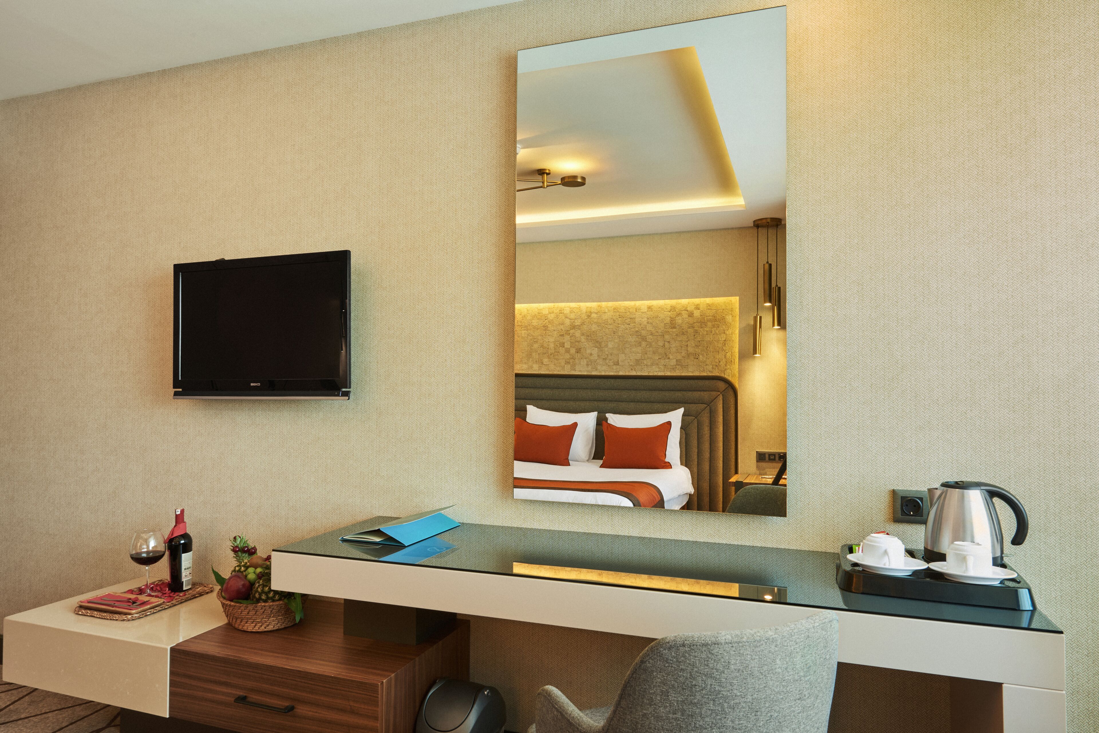 A hotel room with a flat-screen TV, a desk with a basket of fruit, a coffee maker, and a bed with orange pillows.