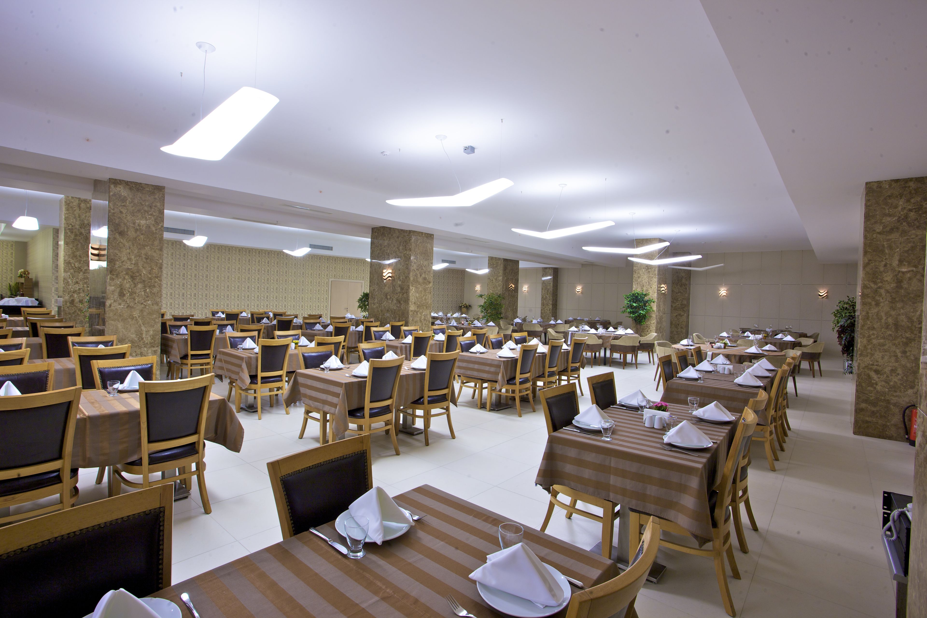 Un comedor amplio con varias mesas y sillas dispuestas en filas, techo alto y lámparas de diseño moderno.