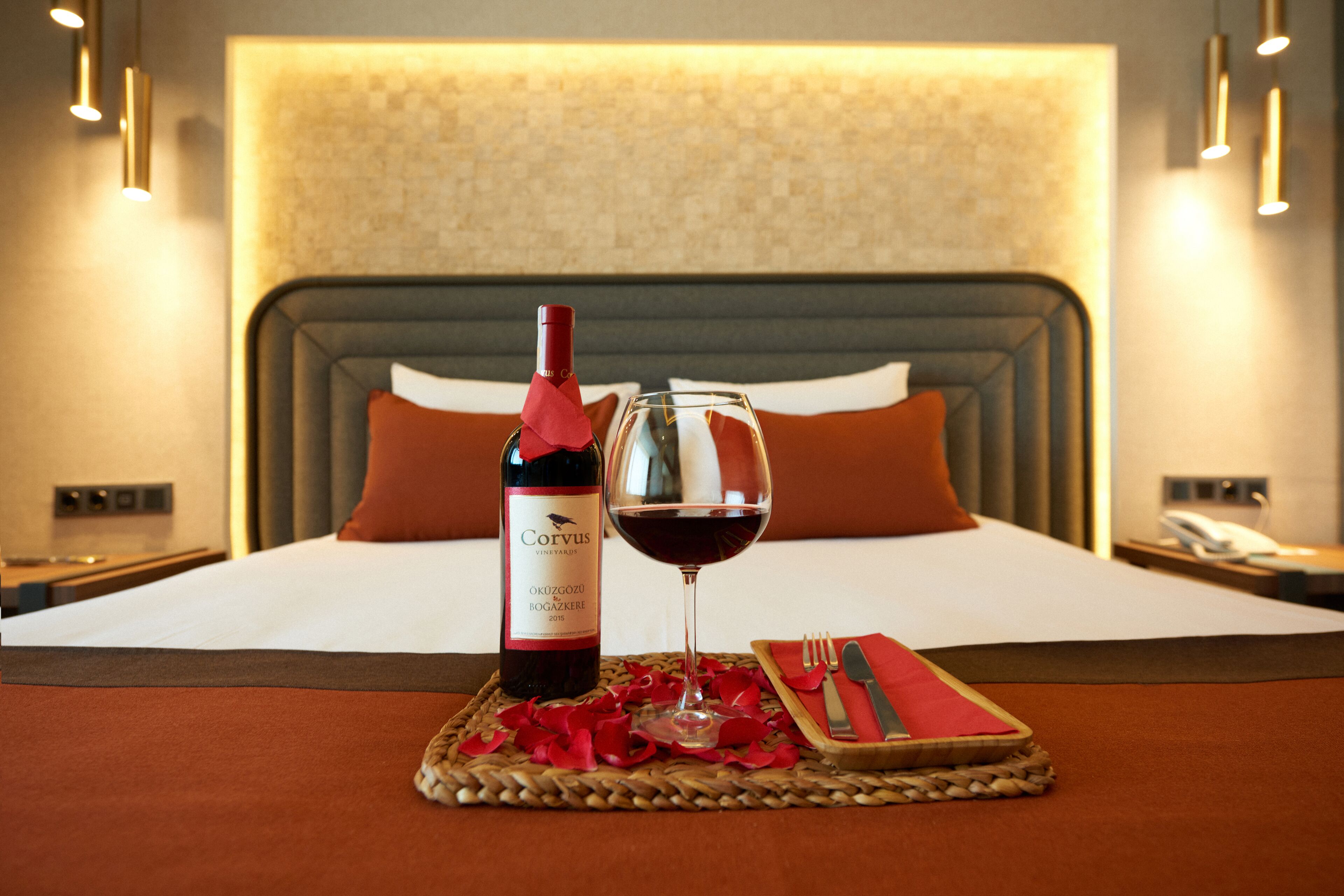 A bed with a headboard, a bottle of wine, a wine glass, and a small table with a napkin and cutlery.