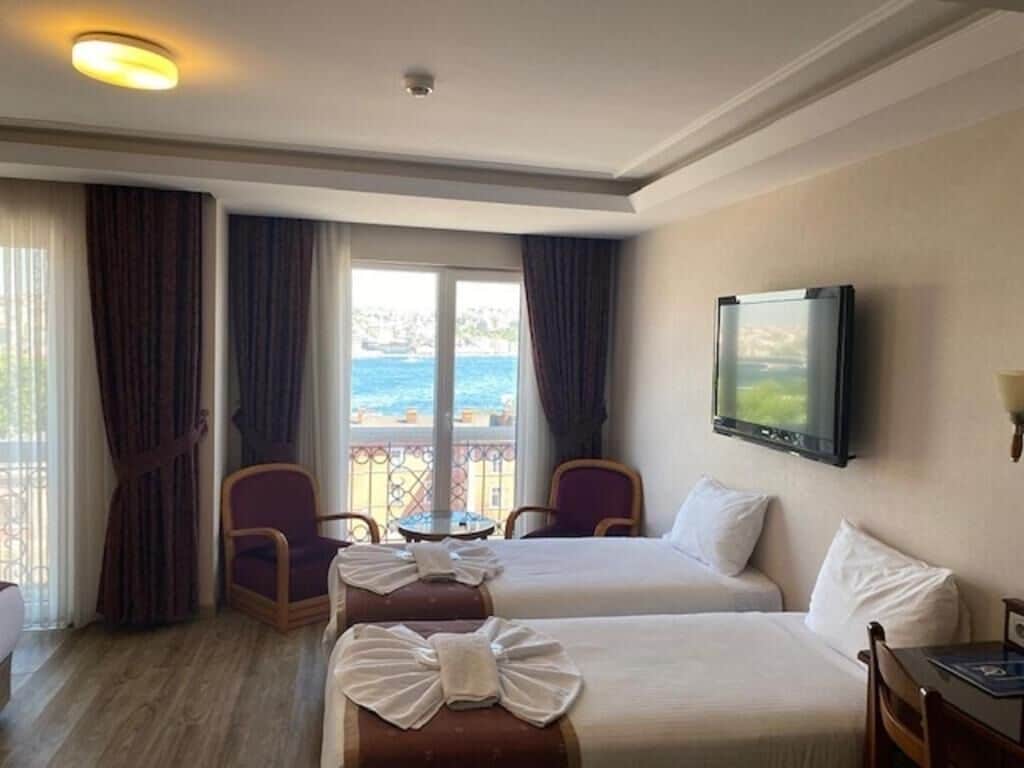A hotel room with two beds, a television, a balcony with a view of the sea, and a table with chairs.