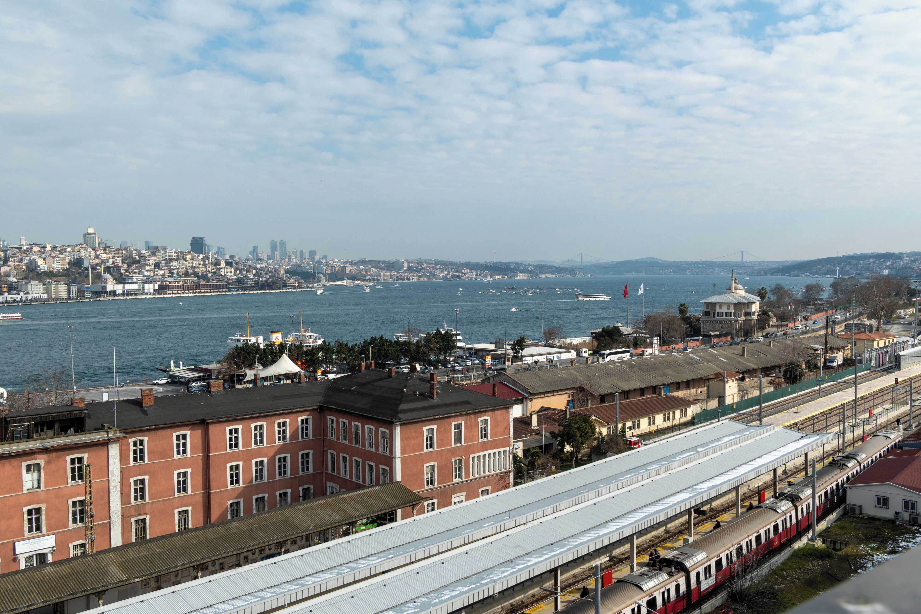 A train station with multiple tracks, a large body of water in the background, and a cityscape across the water.