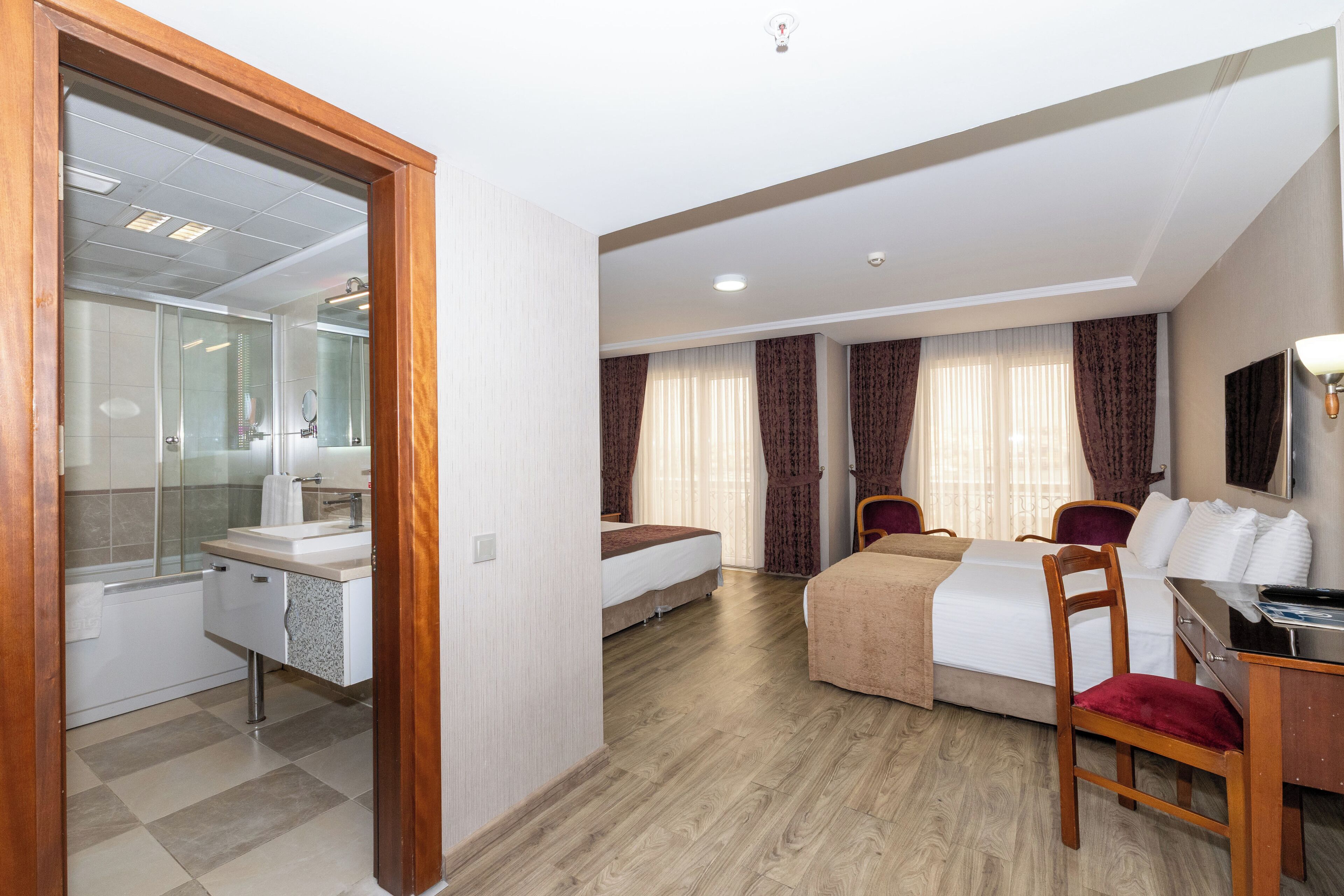 Een hotelkamer met een houten vloer, een bed, een bureau met stoel, een badkamer met spiegel en een raam met gordijnen.