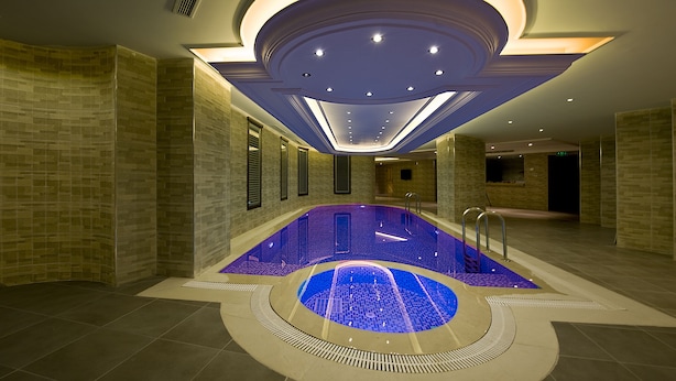 A modern indoor swimming pool with a central blue feature, surrounded by a tiled floor and walls, with recessed lighting and a curved ceiling design. - Askoc Hotel ve Spa (Istanbul)