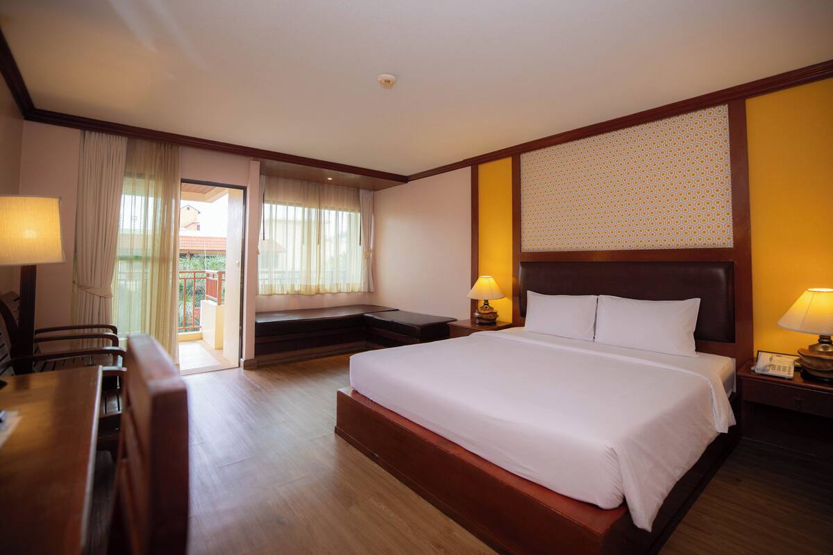 A hotel room with a large bed, a desk, a chair, a nightstand with a lamp, and a view of the outside.