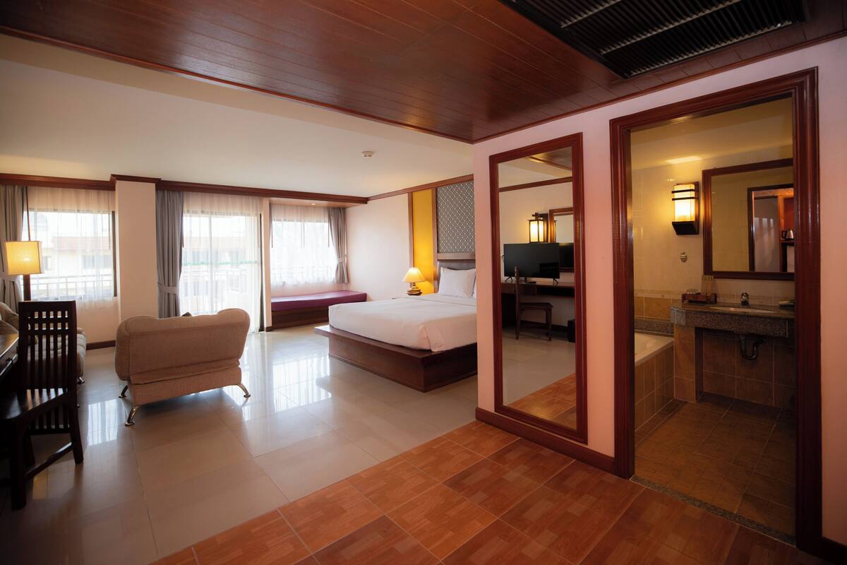 A hotel room with a bed, a desk, a chair, a sofa, a TV, and a bathroom visible through a mirror.