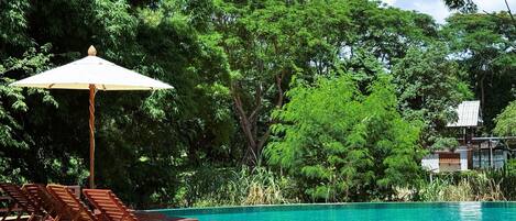 A pool area with wooden lounge chairs and a white umbrella, surrounded by lush greenery and a clear blue sky.
