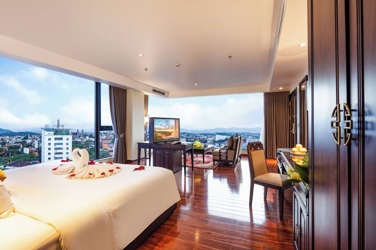A spacious hotel room with a large bed, a desk, a chair, and a view of the city.