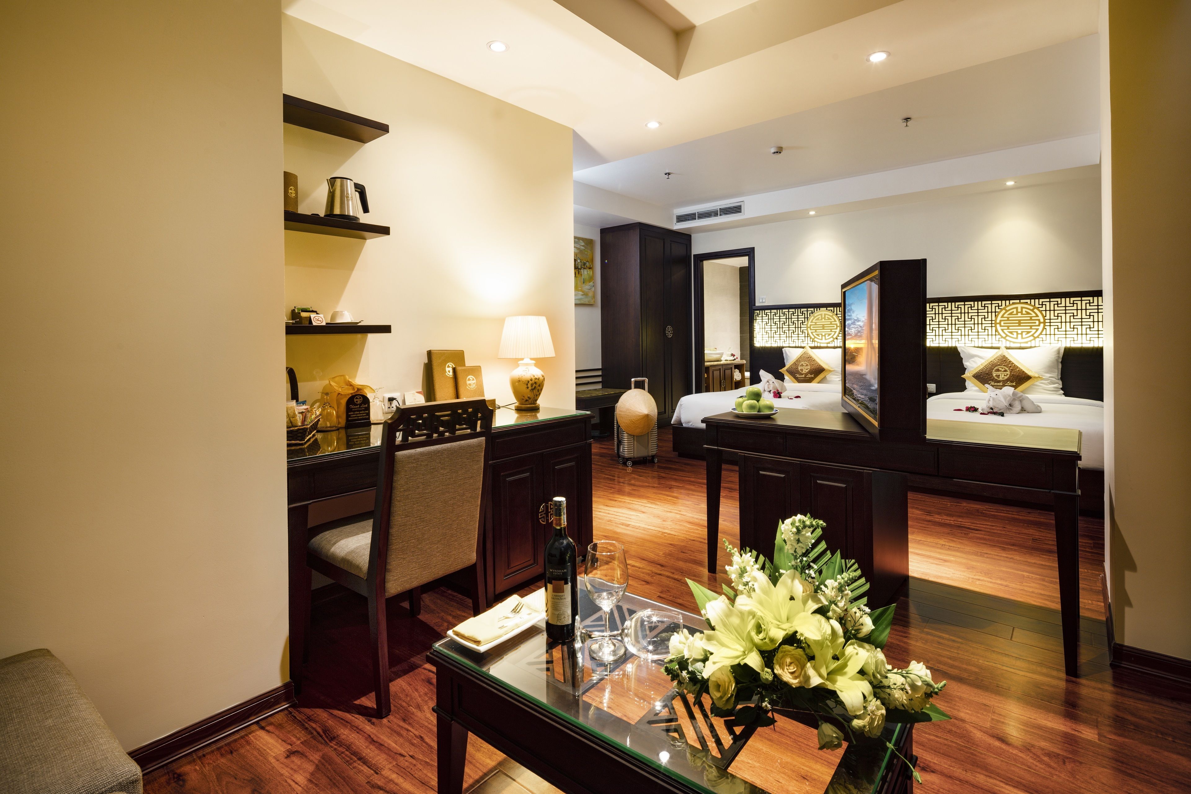 A hotel room with a dining area, a bed with a decorative headboard, a large mirror, and a vase of flowers on the table.