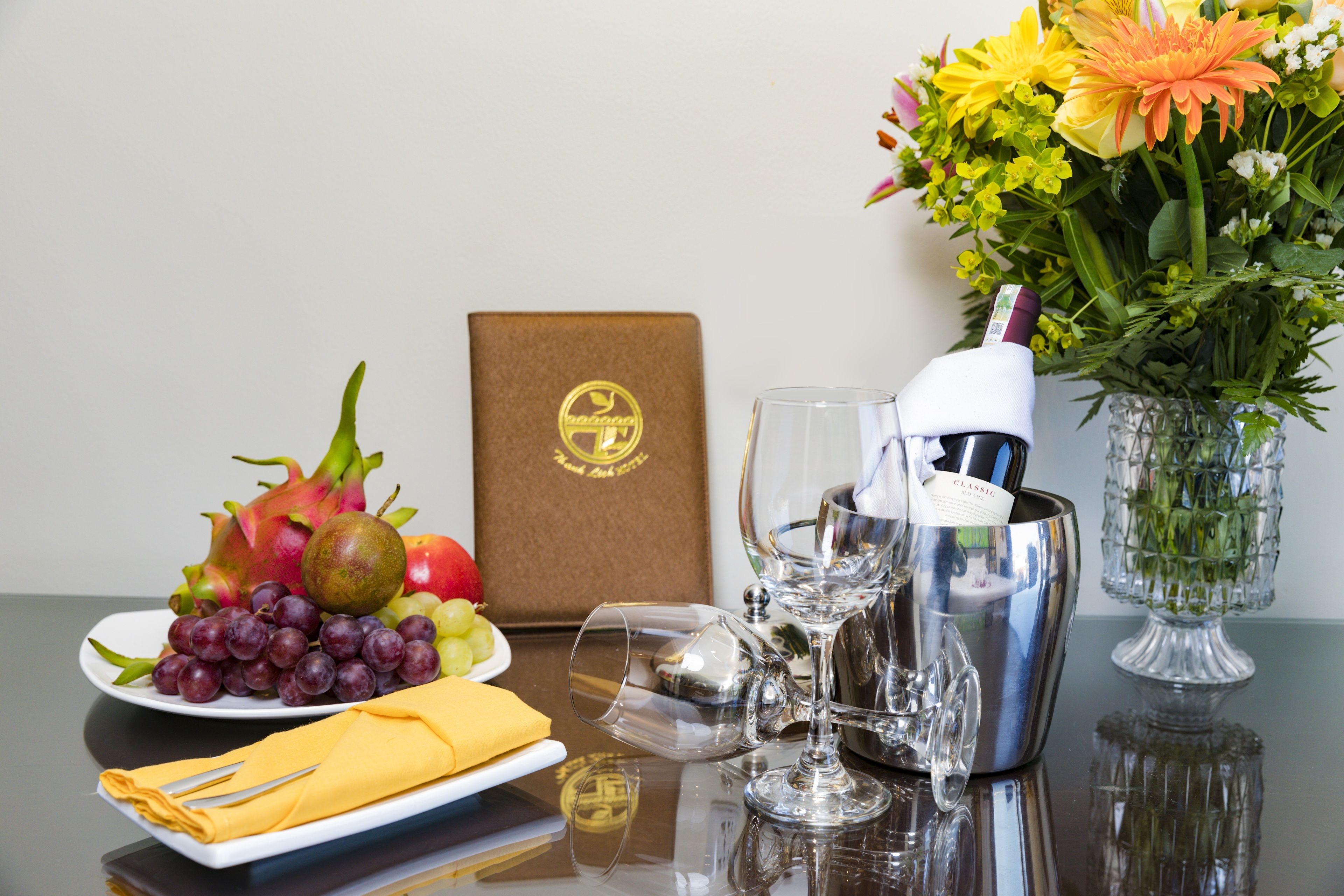 A table with a bottle of champagne, a glass, a bowl, a plate of fruit, a napkin, and a vase with flowers.
