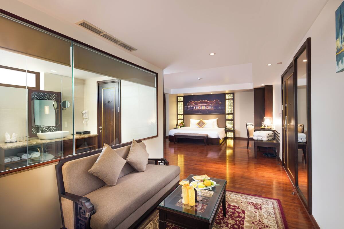A modern hotel room with a sofa, a glass coffee table with fruit, a bed with a nightstand, and a large mirror.