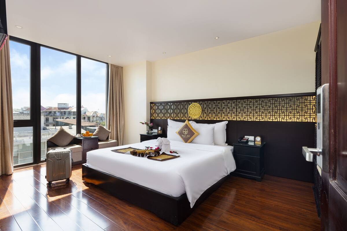 A hotel room with a large bed, a nightstand, a suitcase, and a view of the city through large windows.