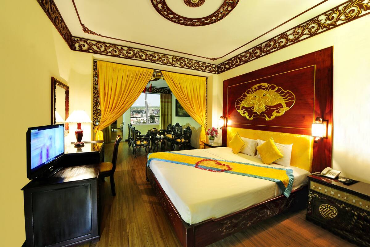 A hotel room with a large bed, a television on a stand, a desk with a lamp, and a view of the city through the curtains.