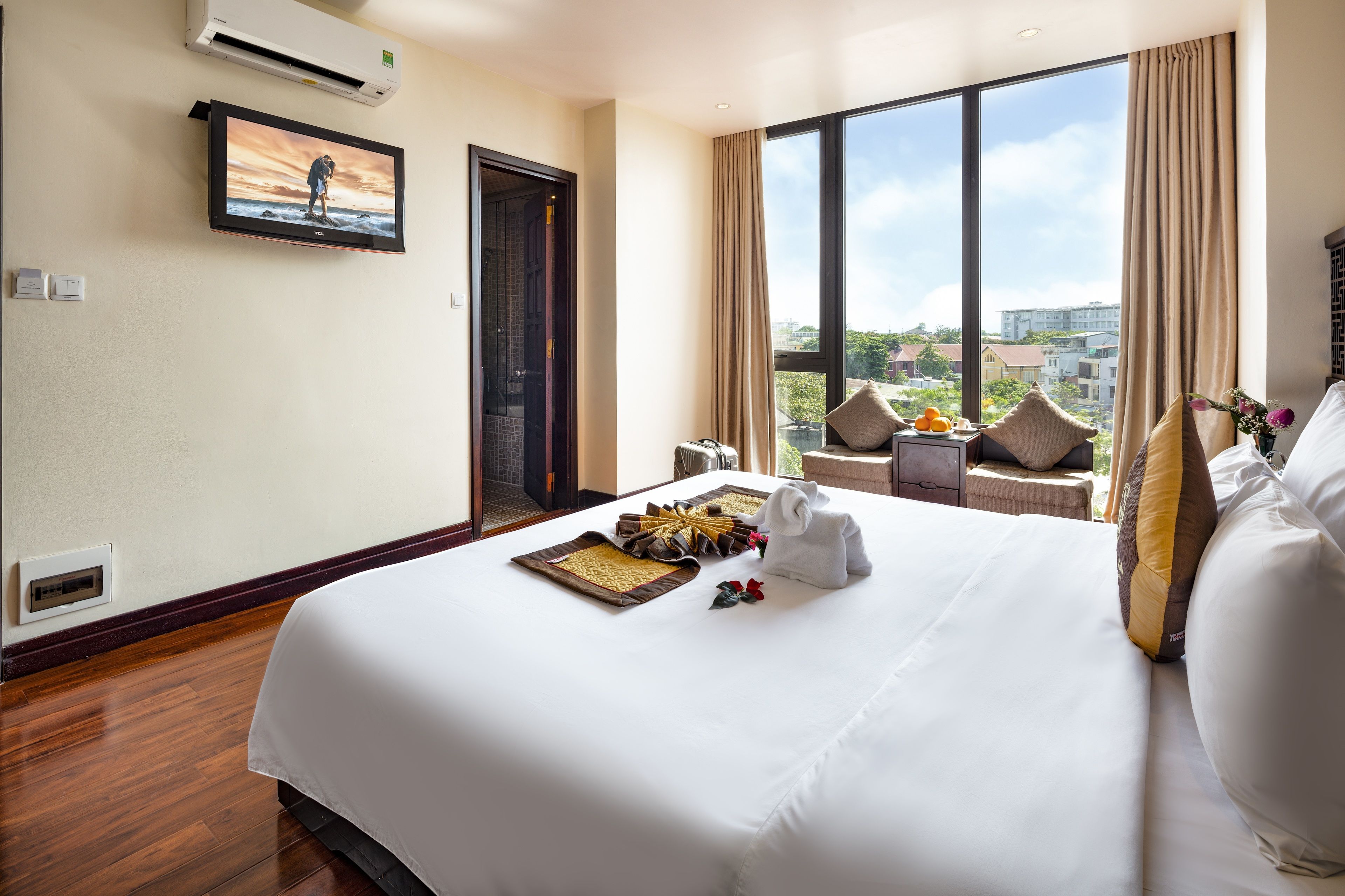 A hotel room with a large bed, a TV on the wall, a view of the city, and a window with curtains.