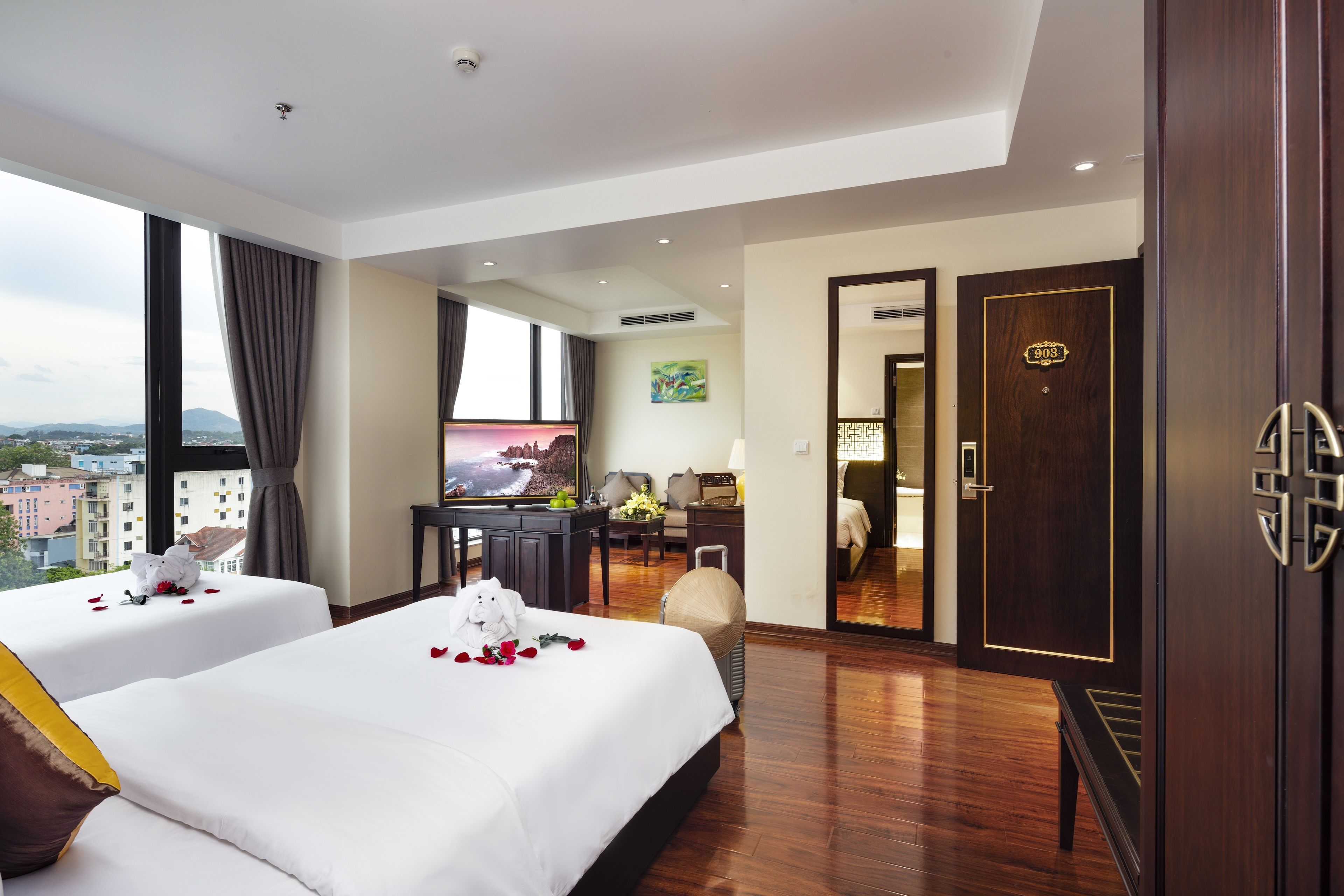 A hotel room with two beds, a desk, a TV, and a view of the city.