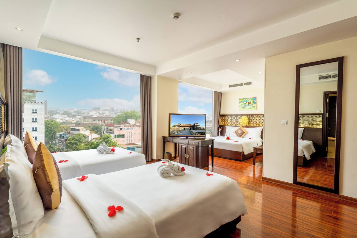 A hotel room with two beds, a desk, a TV, and a large window with a city view.