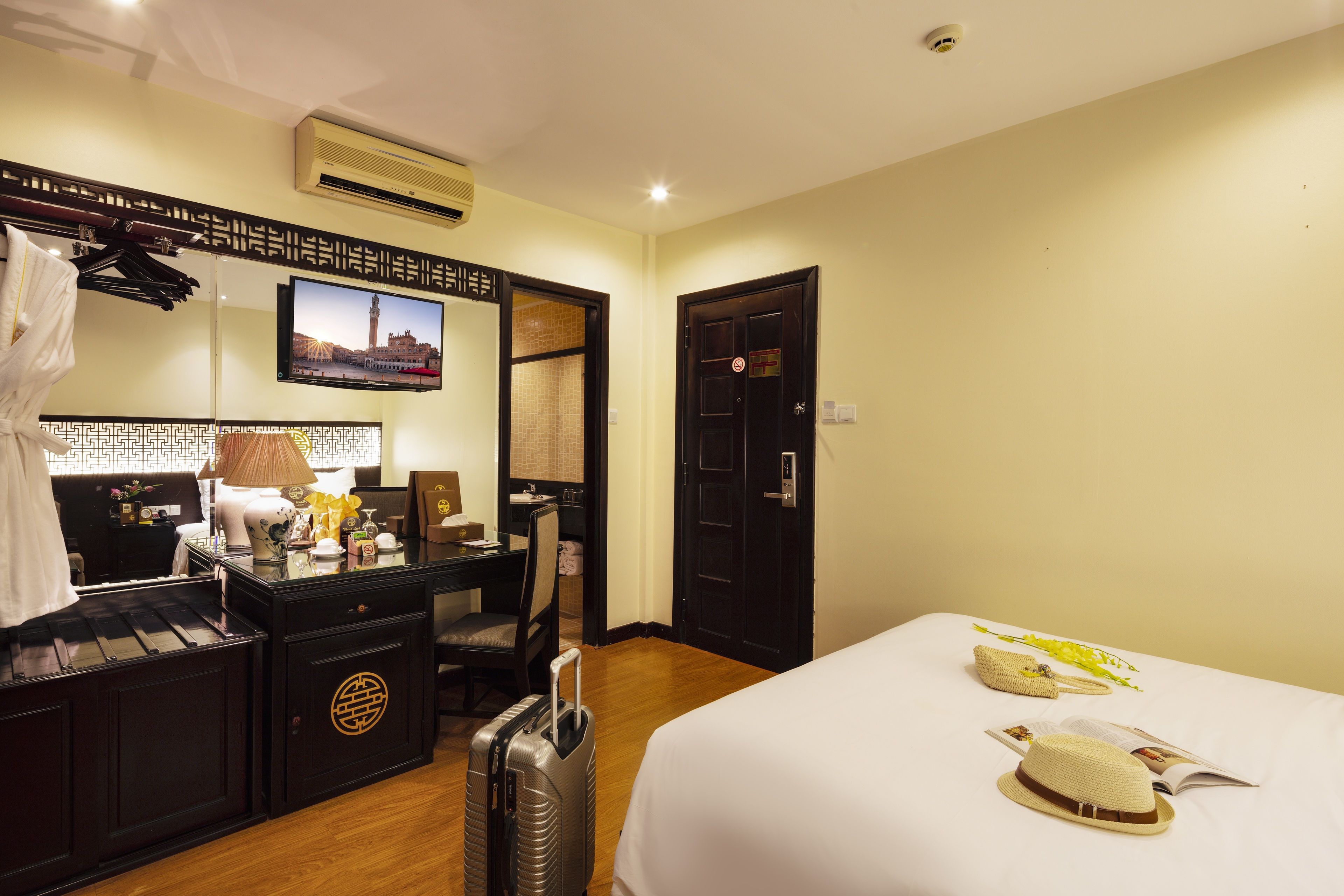 A hotel room with a bed, a desk with a chair, a suitcase, a small table with a lamp, a TV mounted on the wall, and a door.