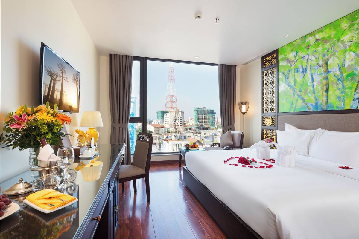 A hotel room with a large bed, a desk with a vase of flowers, a television, and a view of a cityscape with a tower.