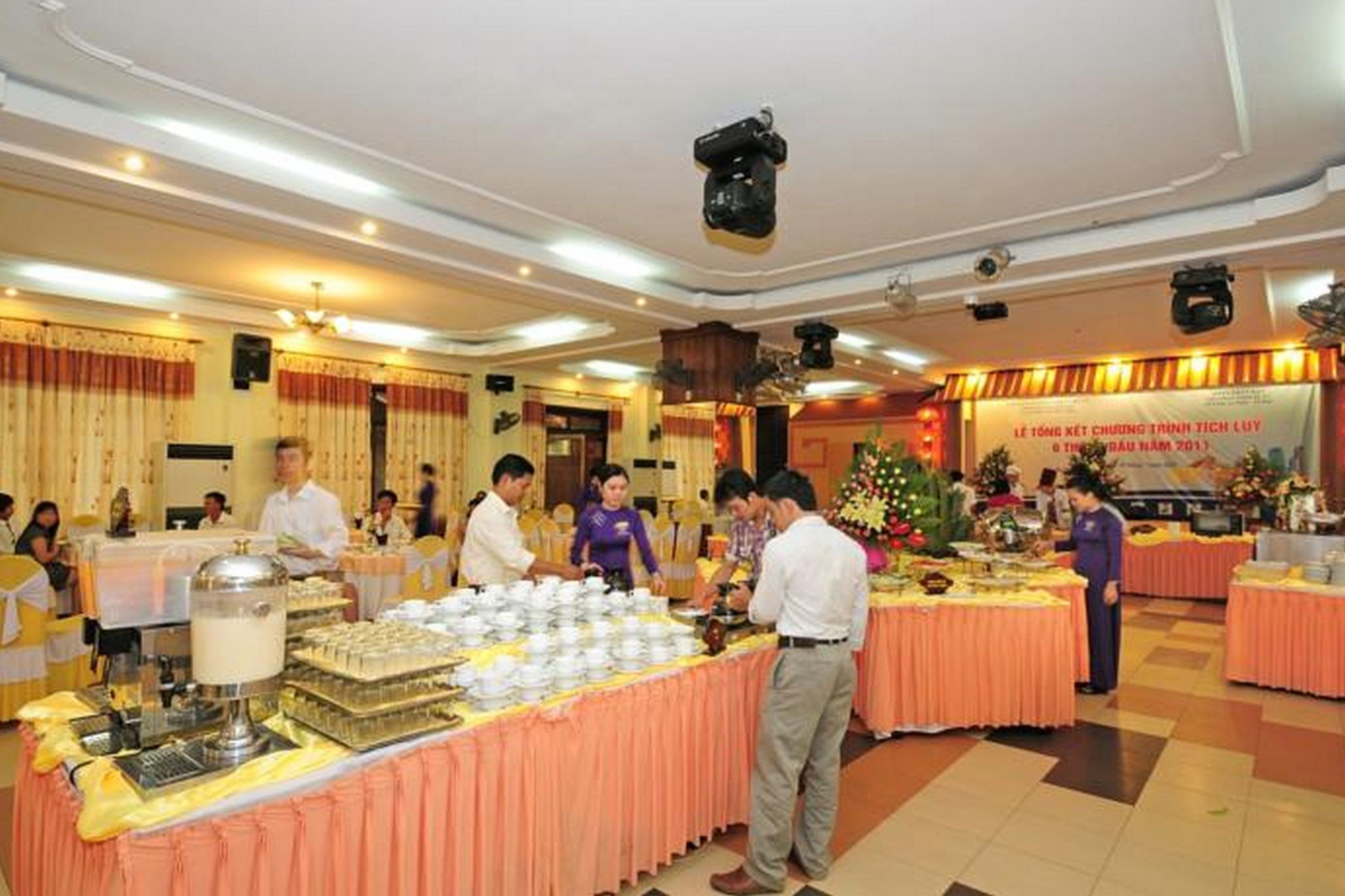 A buffet setup with multiple tables covered in orange tablecloths, people serving and serving food, and a festive atmosphere with decorations.