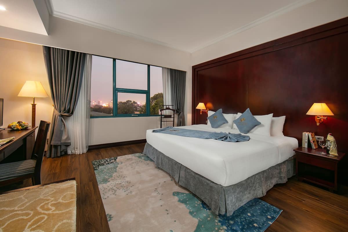 A hotel room with a large bed, two bedside lamps, a desk with a lamp, a chair, and a view of the cityscape through the window.