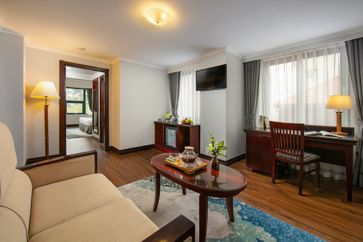 A hotel room with a sofa, a wooden coffee table with a tray, a desk with a chair, a television, and a large window with curtains.