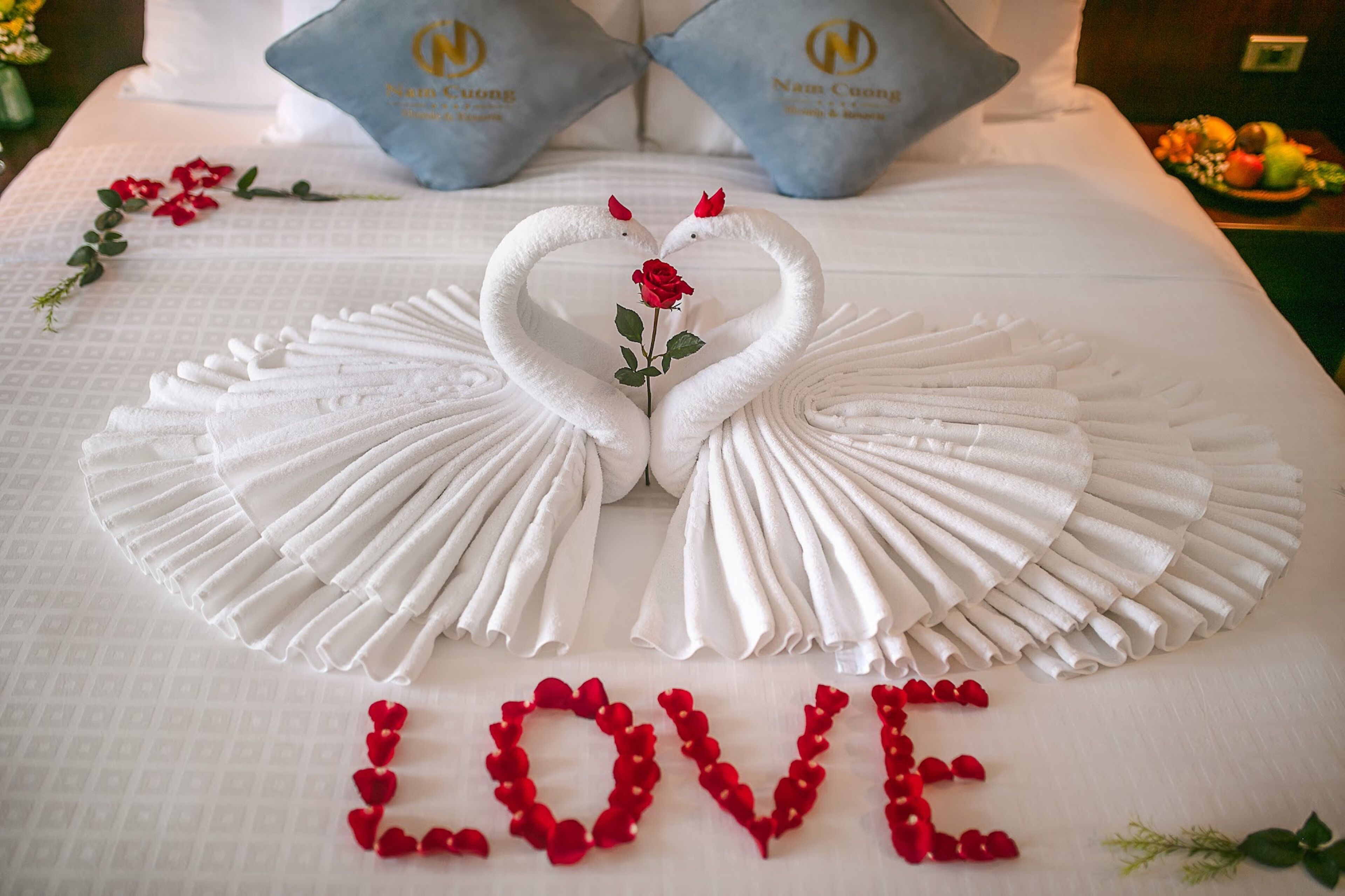 A bed with a towel arrangement in the shape of two swans with a rose in the center, and the word "LOVE" spelled out with rose petals.