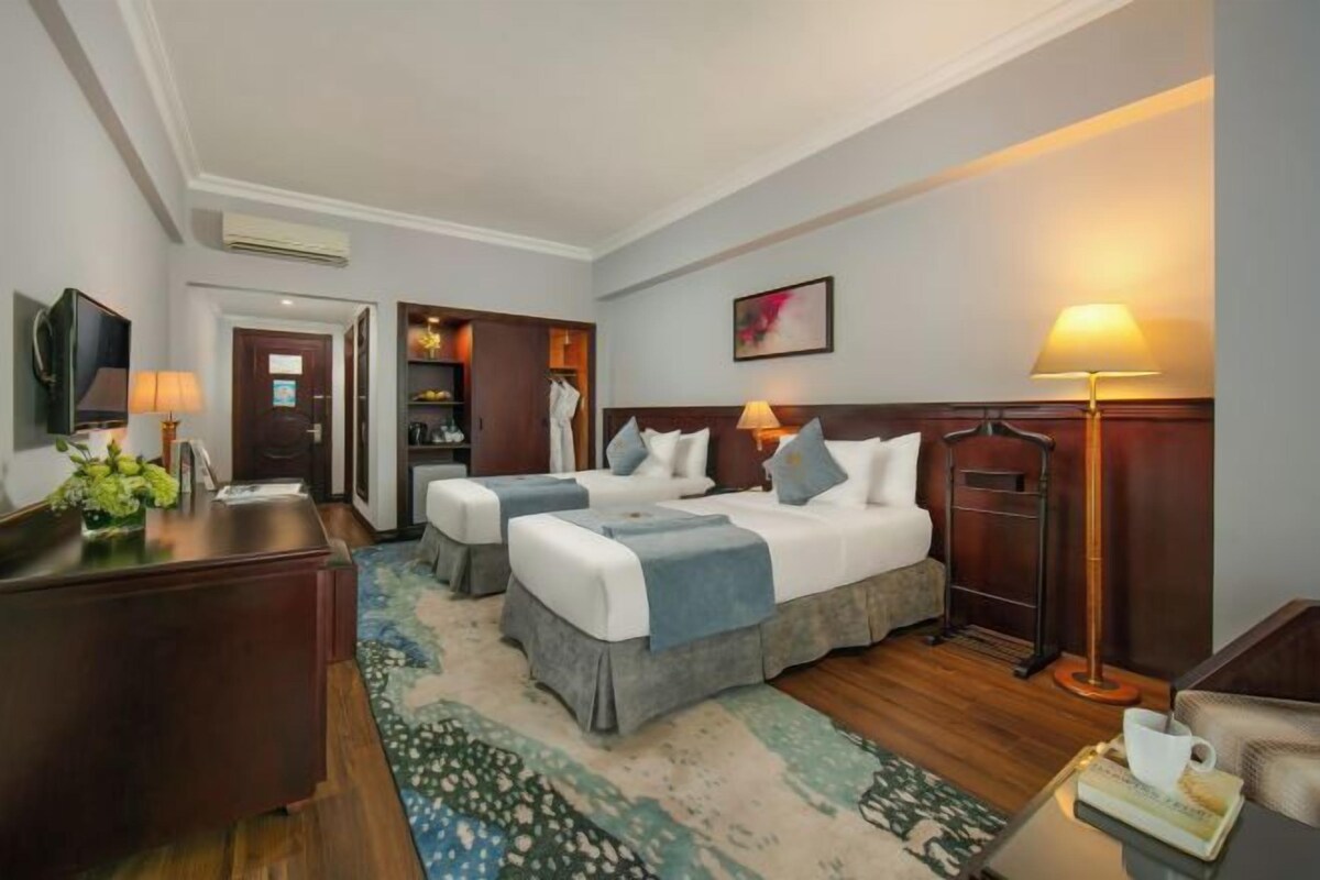 A hotel room with two beds, a desk, a chair, a TV, and a lamp.
