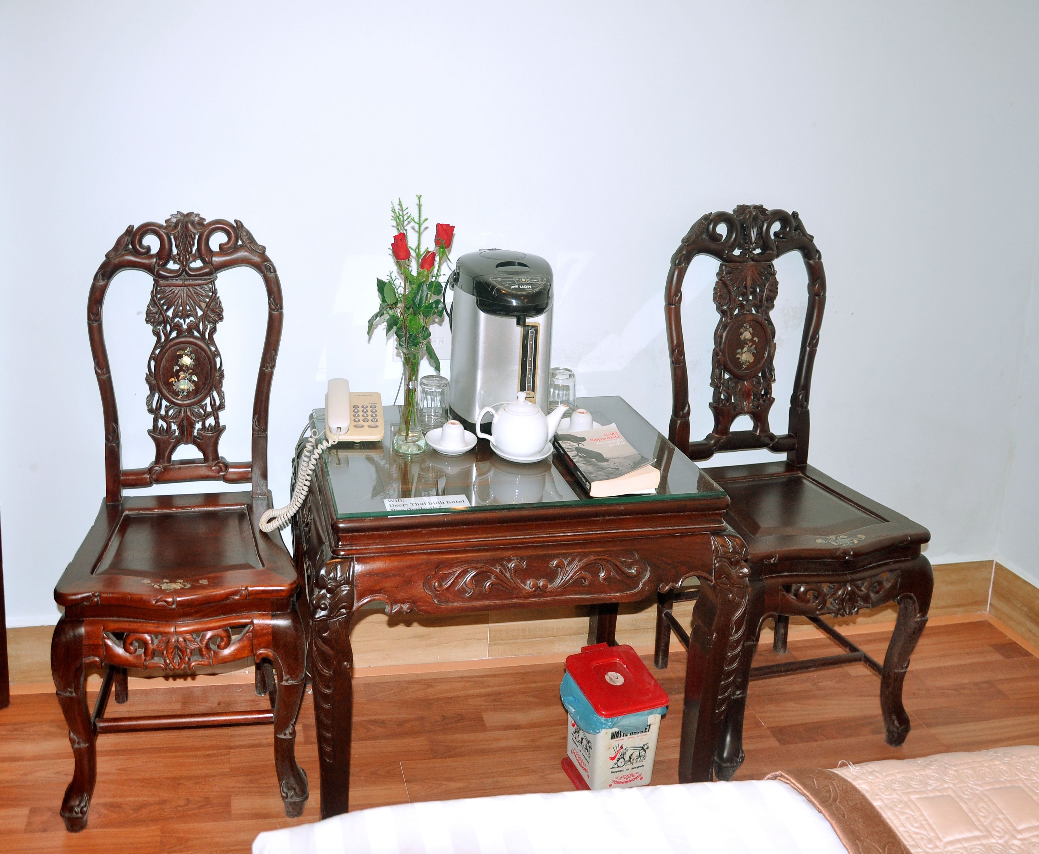 A wooden table with two ornate chairs, a coffee maker, a teapot, a vase with red flowers, and a small trash bin.