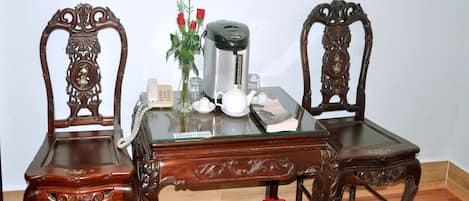 A wooden table with two ornate chairs, a coffee maker, a teapot, a vase with red flowers, and a small trash bin.